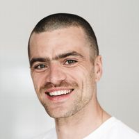 Porträt eines lächelnden Mannes mit kurzem Haar und leichtem Bart, trägt ein weißes T-Shirt. Freundlicher Gesichtsausdruck, natürliche Beleuchtung und unscharfer, heller Hintergrund. Nahaufnahme, die Mimik und Augen betont.