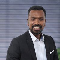Hauptmotiv: Ein lächelnder Mann in dunklem Anzug und weißem Hemd vor unscharfem Bürohintergrund. Porträtaufnahme mit freundlichem Gesichtsausdruck, gepflegtem Bart und einer dezenten Einstecktuch-Detail im Sakkos. Geeignet für professionelle Team- oder Businessdarstellungen.