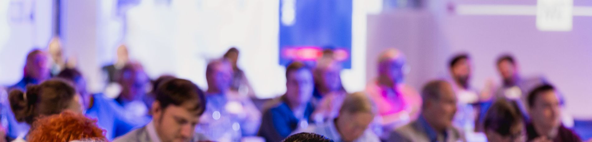 Ein diverses Publikum sitzt in einem Tagungsraum und applaudiert während einer Präsentation; mehrere Reihen mit Teilnehmern, im Vordergrund drei Personen, warme Konferenzatmosphäre.