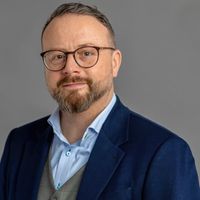 Hochwertiges Business-Kopfporträt eines erwachsenen Mannes mit Brille und kurzem Bart. Er trägt einen dunklen Blazer, ein helles Hemd und eine Weste. Der Hintergrund ist neutral grau, der Blick ist direkt zur Kamera gerichtet und das Gesicht zeigt ein leichtes, freundliches Lächeln. Geeignet für professionelle Profile, Unternehmenskommunikation und Editorials.