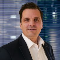 Businessporträt eines lächelnden Mannes im Anzug vor einem Bürohintergrund mit Jalousien und Pflanzen; professionelles Headshot-Foto geeignet für Unternehmensprofile und berufliche Präsentationen.