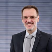 Porträt eines lächelnden Geschäftsmanns mit Brille und grauem Anzug vor einem verschwommenen Bürohintergrund; professionelles Business-Porträt mit freundlichem Ausdruck.
