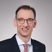 Formelles Porträt eines lächelnden Mannes mittleren Alters als Hauptmotiv. Er trägt einen dunkel gestreiften Anzug, weißes Hemd, rot-weiß gestreifte Krawatte und eine runde Brille. Aufnahme vor neutral hellem Hintergrund in professionellem Business-Stil ohne Identifikation der Person.