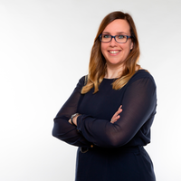 Ein freundliches, professionelles Business-Portrait einer jungen Frau mit Brille, die in einem dunklen Kleid mit verschränkten Armen vor einem hellen Hintergrund in die Kamera lächelt.