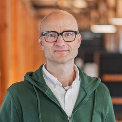 Ein Mann mit Brille und grüner Kapuzenjacke steht in einem modernen Büro mit Holzelementen.