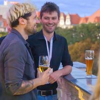 Zwei Männer unterhalten sich bei einem Getränk auf einer Terrasse mit Blick auf die Stadt.