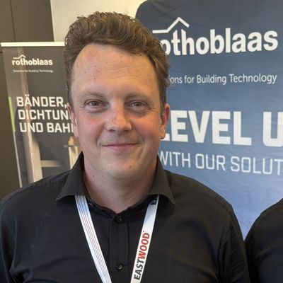 Zwei Männer stehen auf einer Messe vor einem Stand von Rothoblaas. Beide tragen schwarze Hemden und Namensschilder.