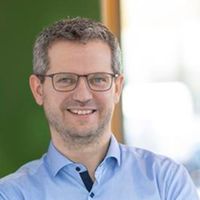 Ein lächelnder Mann mit Brille steht in einem modernen Büro und lehnt an einer Wand.
