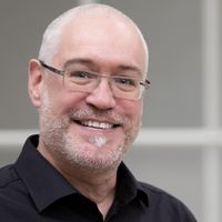 Ein lächelnder Mann mit Brille und Bart in einem schwarzen Hemd.