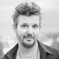 Ein schwarz-weiß Porträt eines Mannes mit lockigem Haar und Bart, der direkt in die Kamera schaut.