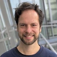 Ein Mann mit dunklem Haar und Bart lächelt in die Kamera, während er vor einem modernen Gebäude steht.