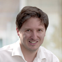 Ein Porträt eines lächelnden Mannes in einem weißen Hemd vor einem unscharfen Hintergrund.