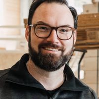 Ein Mann mit Brille und Bart lächelt in die Kamera. Er trägt eine dunkle Jacke und steht in einem modernen Innenraum.