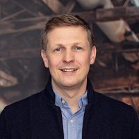 Ein Mann steht in einer verlassenen Fabrik mit sichtbaren Stahlträgern und Rohren im Hintergrund.