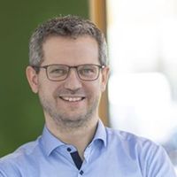 Ein lächelnder Mann mit Brille steht in einem modernen Büro und lehnt an einer Wand.