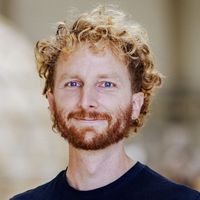 Ein Mann mit lockigem Haar und Bart steht in einem Innenraum und trägt ein schwarzes T-Shirt.