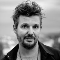 Ein schwarz-weiß Porträt eines Mannes mit lockigem Haar und Bart, der direkt in die Kamera schaut.