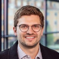 Ein Mann mit Brille und Bart lächelt in die Kamera. Er trägt ein gestreiftes Hemd und eine dunkle Jacke. Im Hintergrund sind unscharfe Fenster zu sehen.