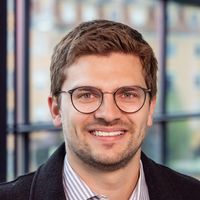 Ein Mann mit Brille und Bart lächelt in die Kamera. Er trägt ein gestreiftes Hemd und eine dunkle Jacke. Im Hintergrund sind unscharfe Fenster zu sehen.