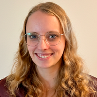 Ein freundliches Porträt einer jungen Frau mit langen, blonden, welligen Haaren und einer modernen Brille, die vor einem hellen Hintergrund in die Kamera lächelt.