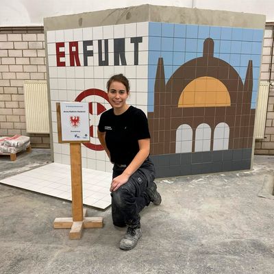 Eine Fliesenlegerin kniet vor einem Mosaik, das den Schriftzug "Erfurt" und ein stilisiertes Gebäude zeigt.