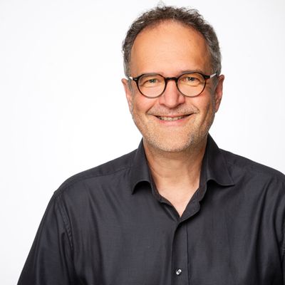 Ein Porträt eines lächelnden Mannes mit Brille und grauem Hemd vor einem weißen Hintergrund.