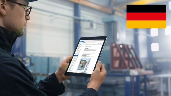 Ein Arbeiter in einer Fabrik hält ein Tablet in der Hand, während er auf den Bildschirm schaut. Im Hintergrund sind Maschinen zu sehen.