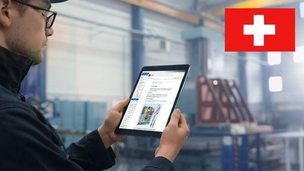 Ein Arbeiter in einer Fabrik hält ein Tablet, während er eine technische Zeichnung betrachtet. Im Hintergrund ist eine Produktionshalle zu sehen.