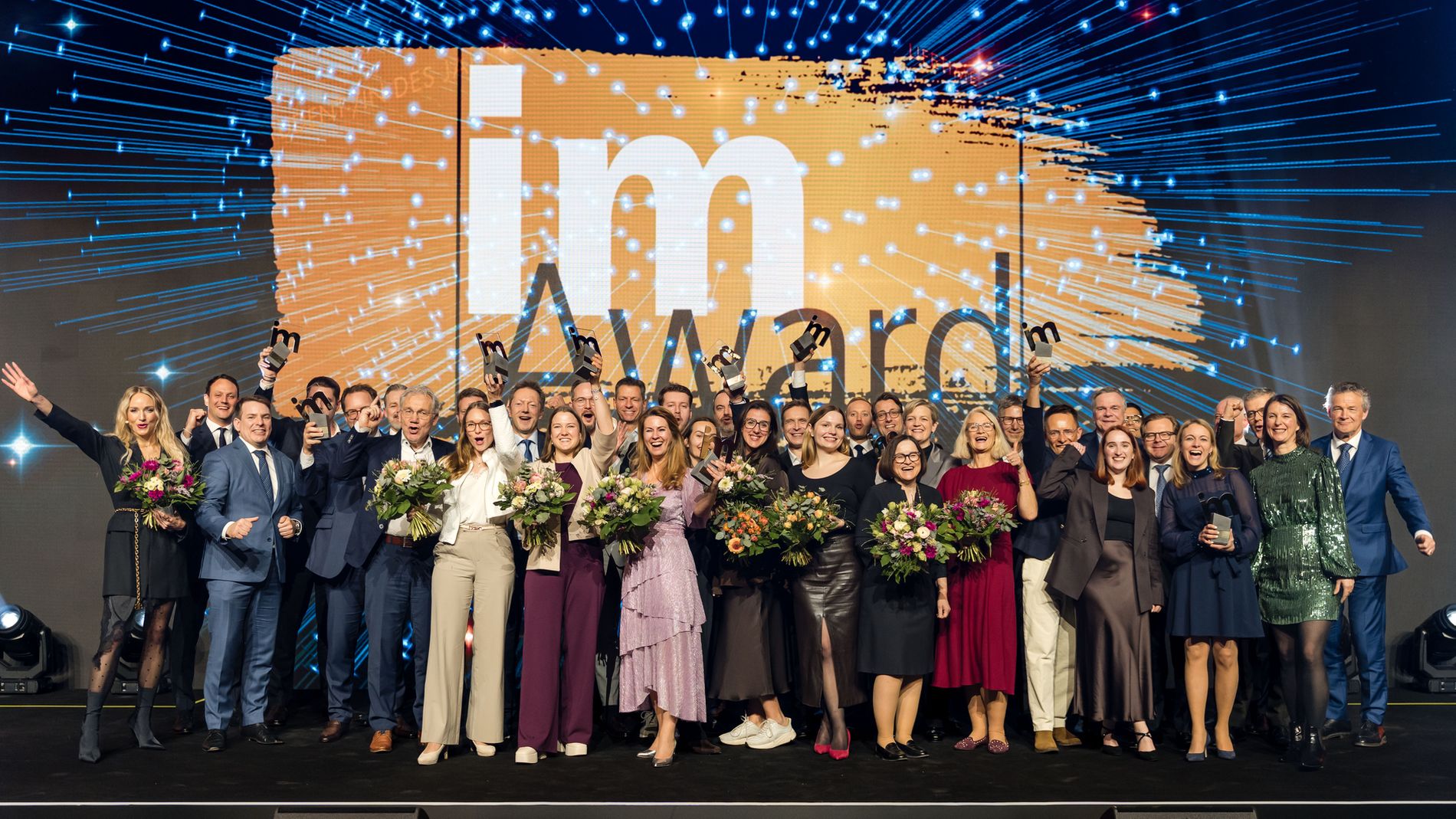 Großes Gruppenfoto von Preisträgern auf einer Bühne bei einer feierlichen Award-Verleihung; die Personen halten Trophäen und Blumensträuße, sitzen in festlicher Kleidung vor einer leuchtenden Medienwand.